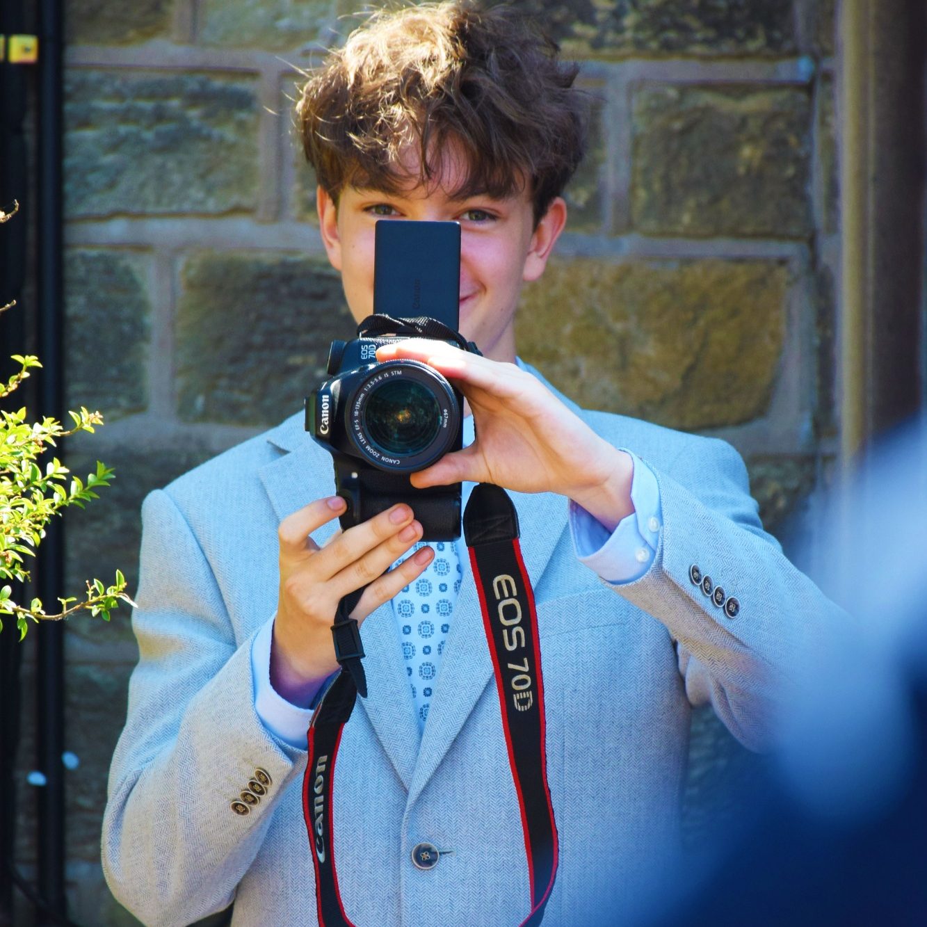 A young man staring at a camera holding up a professional camera in a white and red tshirt